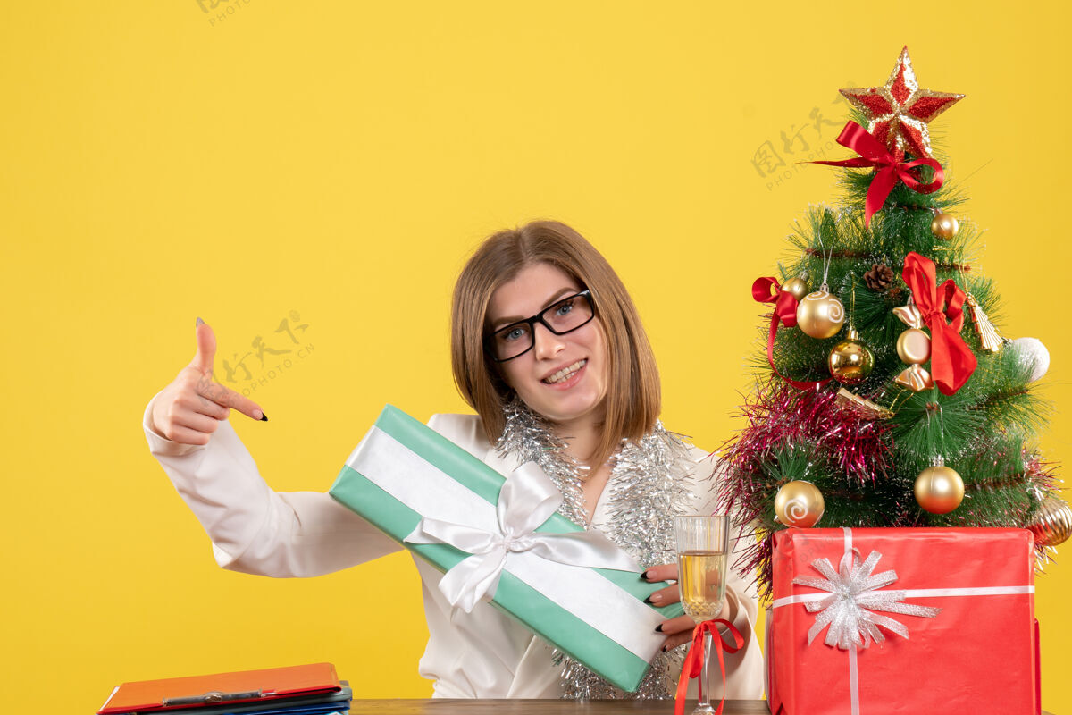 成人正面图：女医生坐在桌子前面 黄色背景上有礼物和树背景微笑礼物