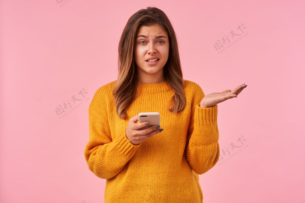 站立室内画像年轻的棕色头发困惑的女性与不高兴的脸和提高困惑的手 而站在粉红色的智能手机棕色女性表情
