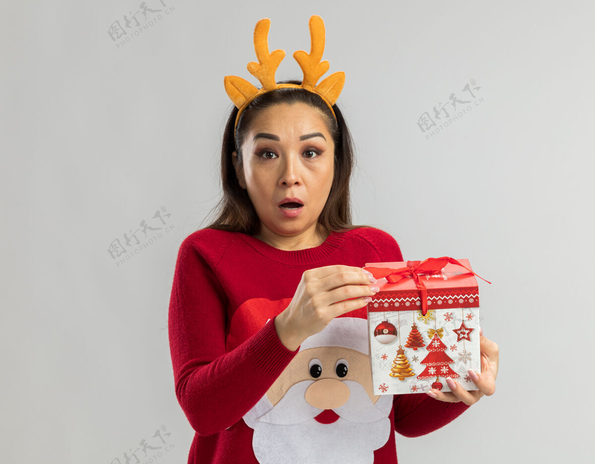 边穿着红色圣诞毛衣的年轻女子 戴着滑稽的鹿角边 手里拿着圣诞礼物 看起来很惊讶拿着有趣站着