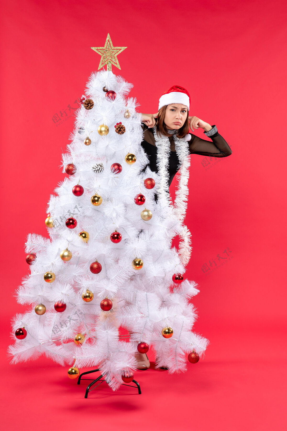 人物圣诞心情与年轻漂亮的女士在一个黑色的礼服和圣诞老人的帽子藏在新年树后面心情雪人帽子