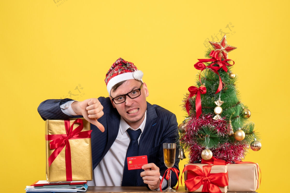 喜剧演员前视图的商人与圣诞树附近的桌子上坐着的圣诞帽制作拇指向下的标志和黄色的礼物向下男人生意人