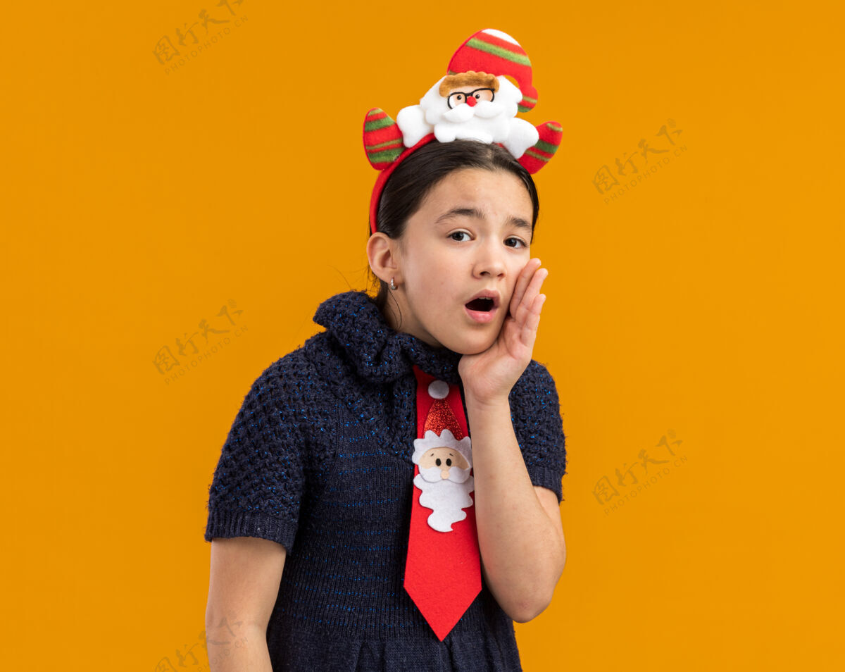 头小女孩穿着针织连衣裙 头上系着一条红色领带 戴着一个有趣的圣诞戒指 手靠着嘴低语着新年新衣服