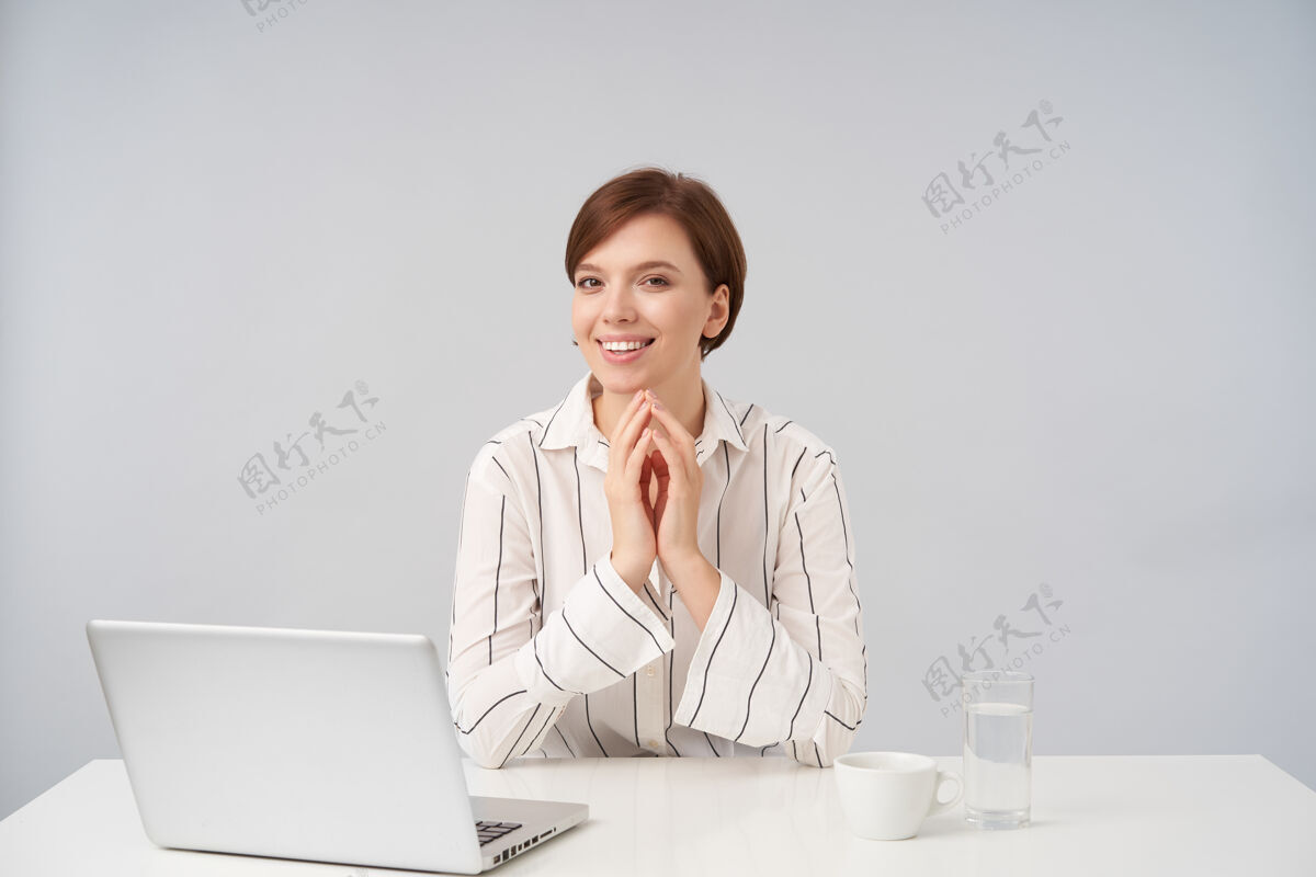 心情快乐的棕色头发的年轻女子 自然化妆 双手合十 面带迷人的笑容 坐在白色的椅子上工作好坐着