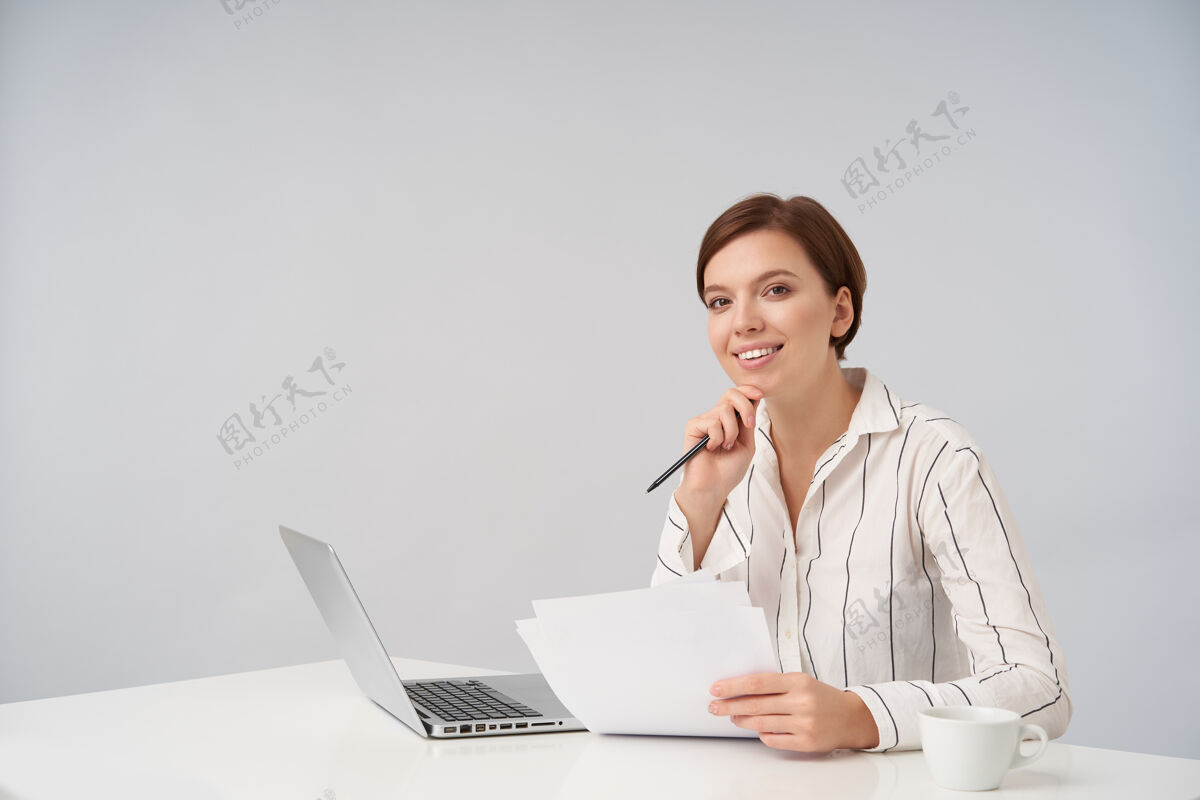 室内快乐的年轻棕色眼睛短发女子愉快地微笑着 下巴靠在抬起的手上 拿着笔记本电脑和文件摆在白色的桌子上女商人坐着条纹
