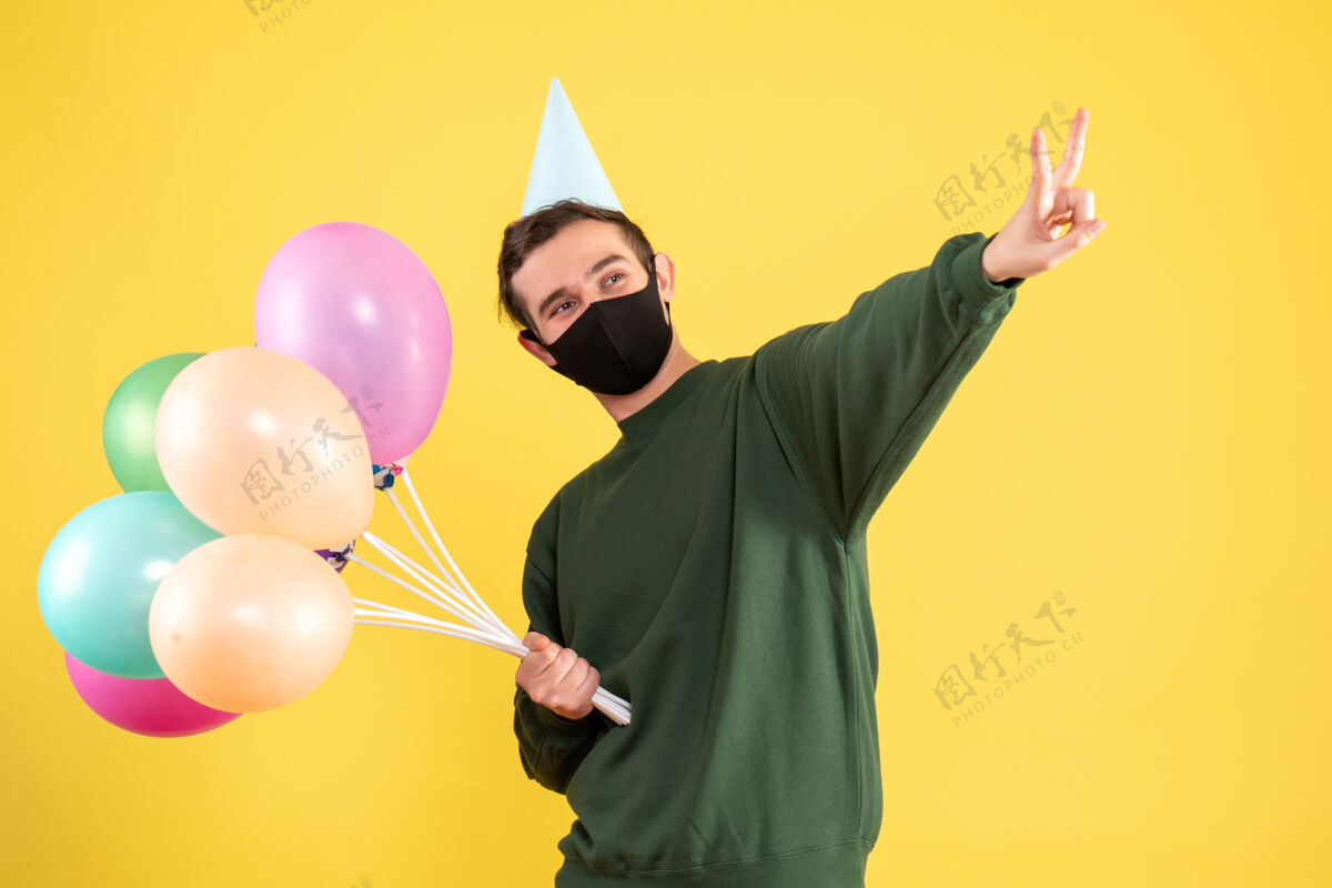 气球正面图：戴着党帽的年轻人和五颜六色的气球站在黄色的墙上做着胜利的标志站着年轻人五颜六色