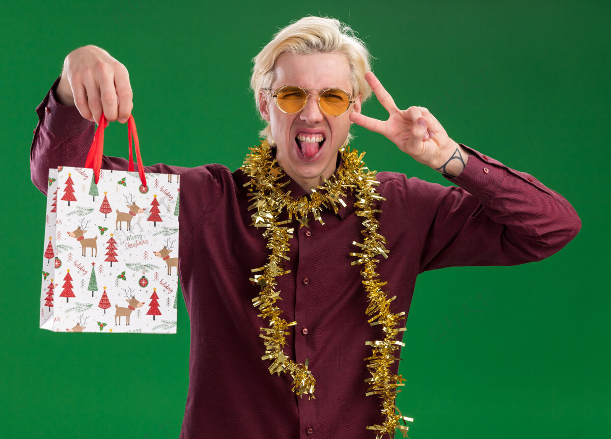 圣诞节顽皮的年轻金发男子戴着眼镜 脖子上戴着金属丝花环 手里拿着圣诞礼品袋 看着相机 露出舌头 做着绿色背景上孤立的和平手势和平金发金箔