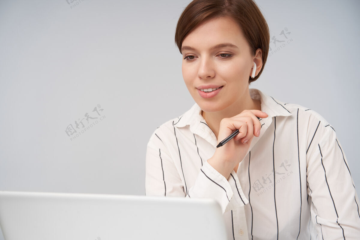 年轻一位年轻漂亮的短发黑发女士的肖像 她剪着时髦的发型 举起手中的笔 面带微笑 看着笔记本电脑的屏幕 摆出白色的姿势模特姿势棕色