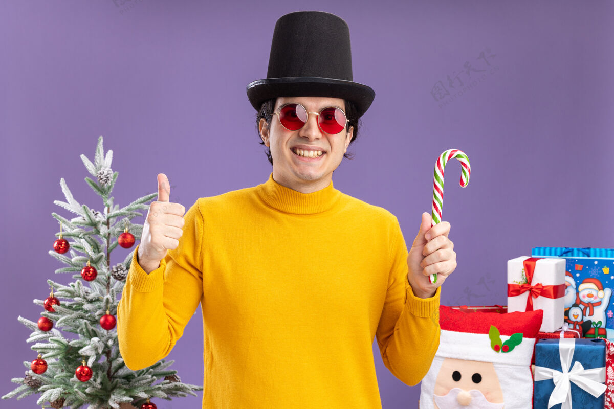 圣诞节穿着黄色高领毛衣戴着眼镜戴着黑色帽子拿着糖果手杖微笑着竖起大拇指站在一棵圣诞树旁紫色墙上的礼物男人糖果高领毛衣