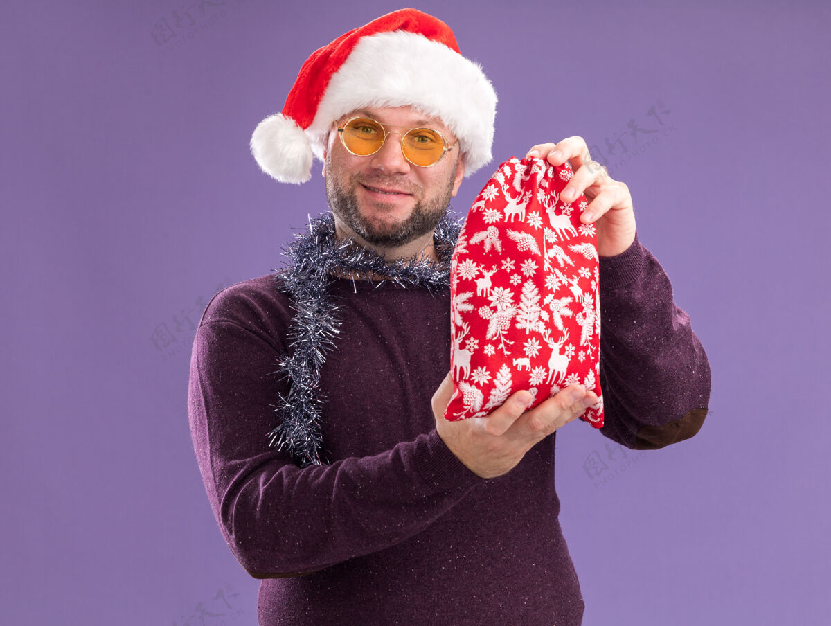 脖子高兴的中年男子戴着圣诞帽 脖子上戴着金箔花环 戴着眼镜 手里拿着圣诞礼品袋 隔离在紫色的墙上围着戴着麻袋