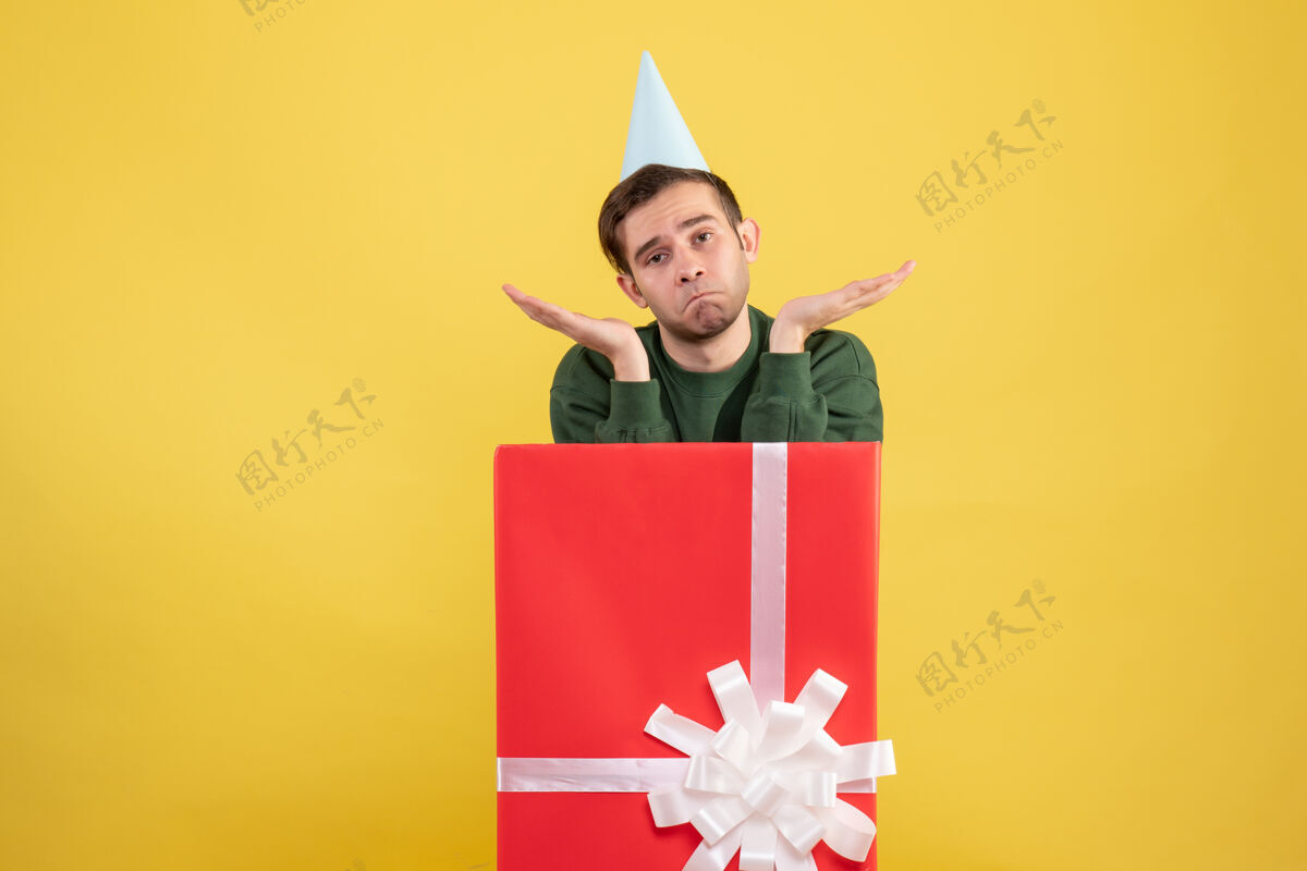 礼物前视图迷惑了一个戴着党帽的年轻人站在黄色的大礼盒后面地点礼品盒大
