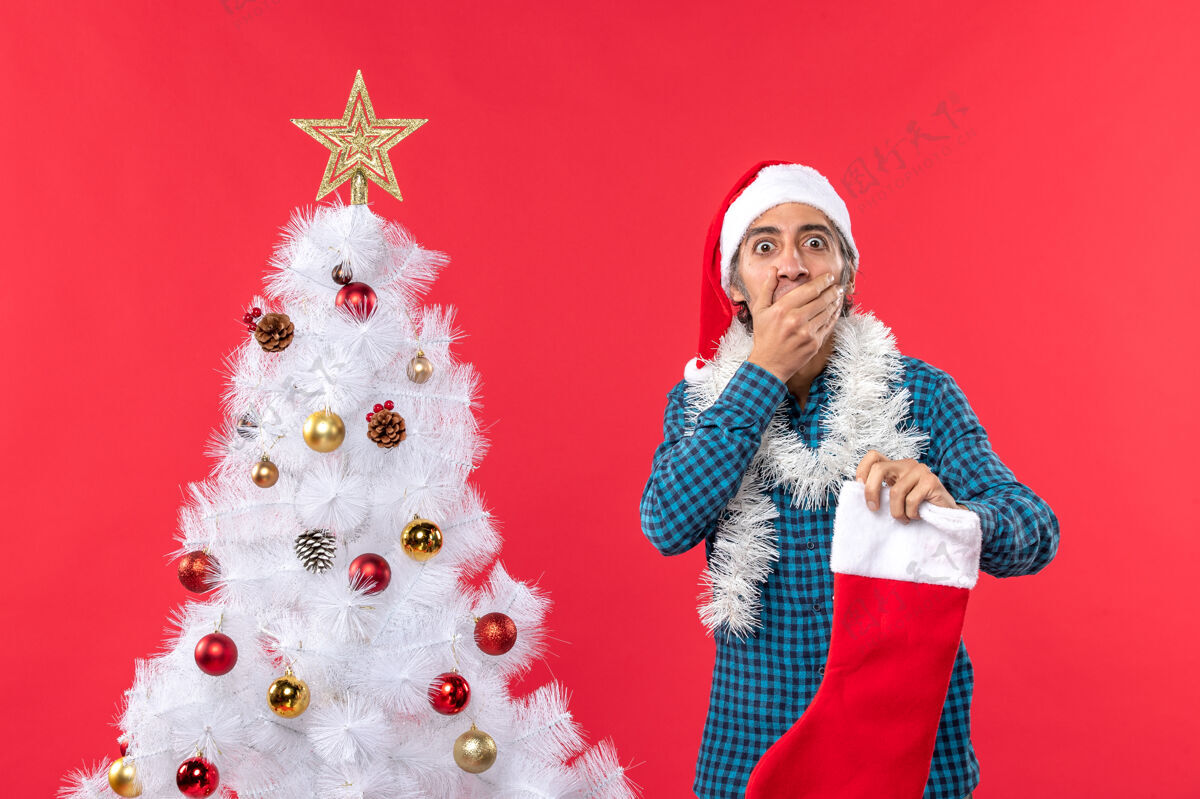 帽子一个戴着圣诞老人帽子 穿着蓝色条纹衬衫 手里拿着圣诞袜子的年轻人闭上了嘴震惊圣诞老人男人