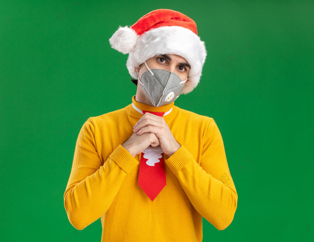 帽子穿着黄色高领毛衣 戴着圣诞老人帽 打着滑稽领带 戴着面罩 手牵着手站在绿色背景下看着摄像机年轻高领毛衣面具