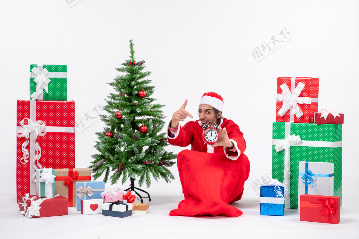 展示年轻人坐在地上庆祝圣诞节 并在礼物和圣诞树旁展示钟表年轻人地树