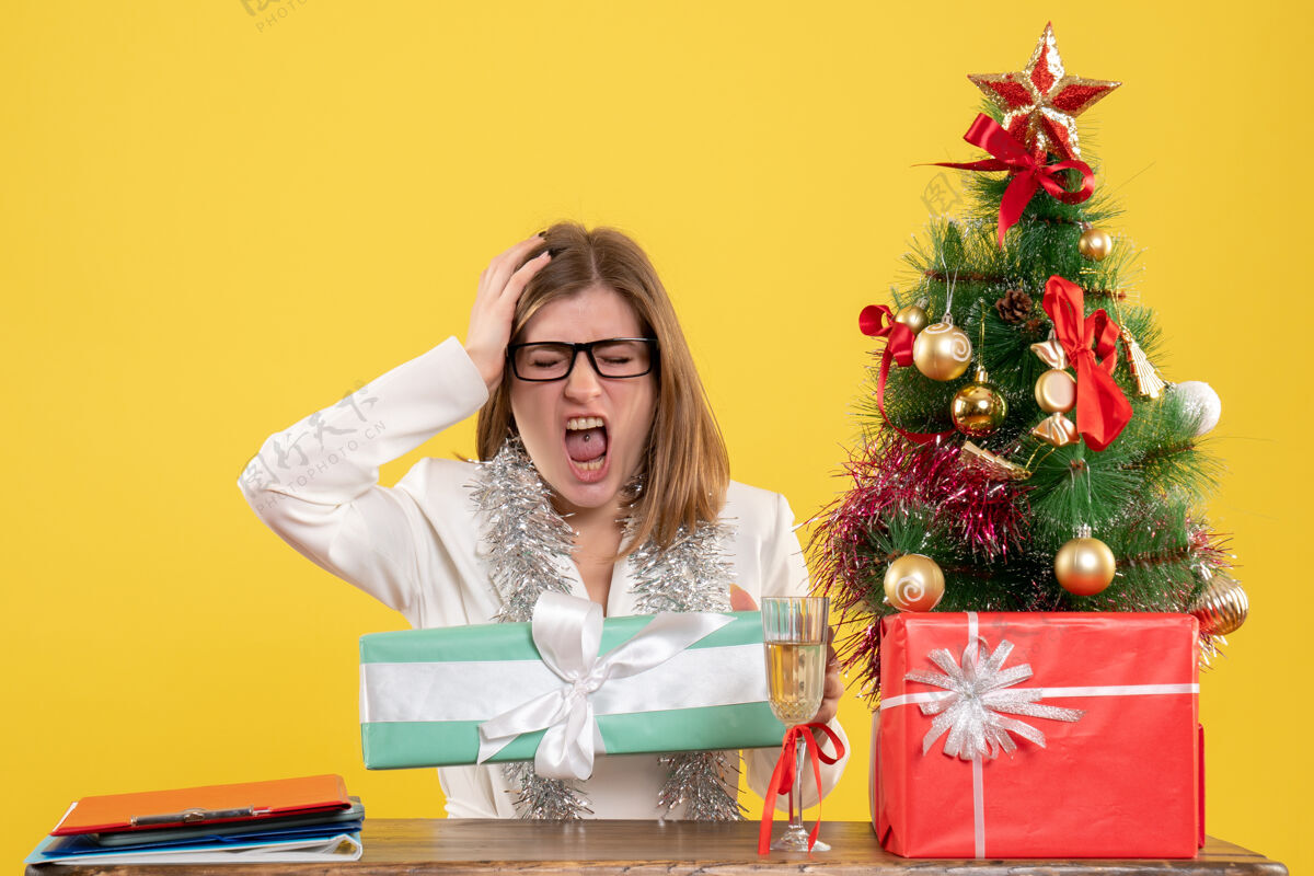 树前视图女医生坐在桌子前 黄色背景上有礼物和圣诞树人礼物女性