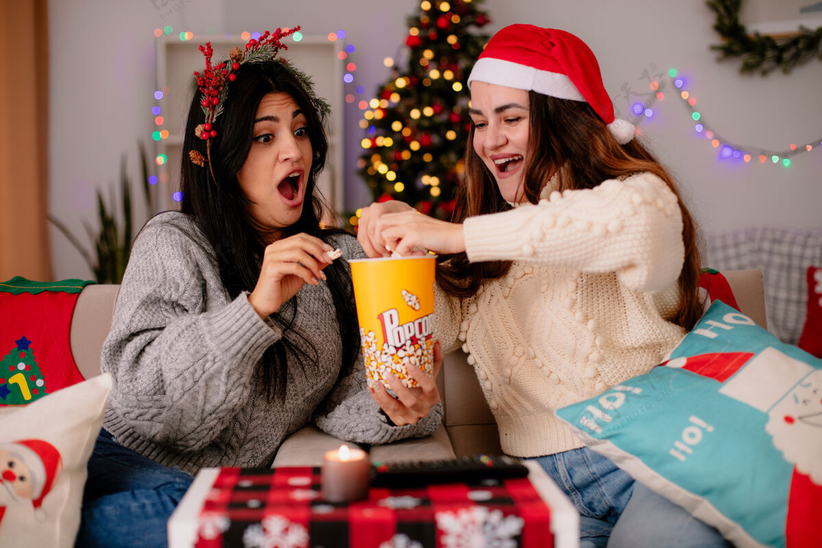 享受带着圣诞帽和冬青花环的年轻女孩们兴奋地抱着爆米花桶 坐在扶手椅上 在家里享受圣诞节时光圣诞快乐坐兴奋