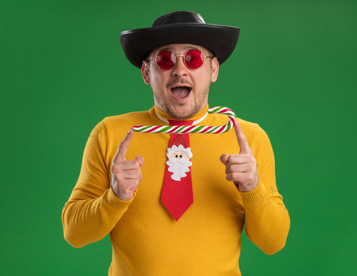 帽子一个穿着黄色高领毛衣 戴着眼镜 打着有趣的红领带 戴着黑色帽子 手里拿着糖果棒的年轻人站在绿色的墙上 高兴而又惊讶圣诞节年轻男人