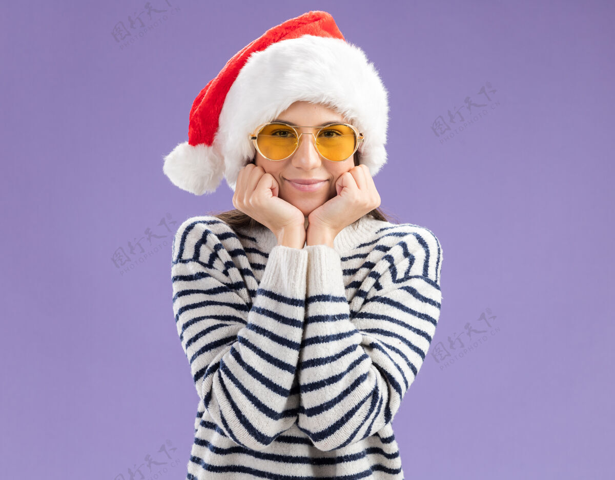 年轻戴着太阳眼镜戴着圣诞帽的年轻白种女孩高兴地把手放在脸上圣诞快乐脸太阳