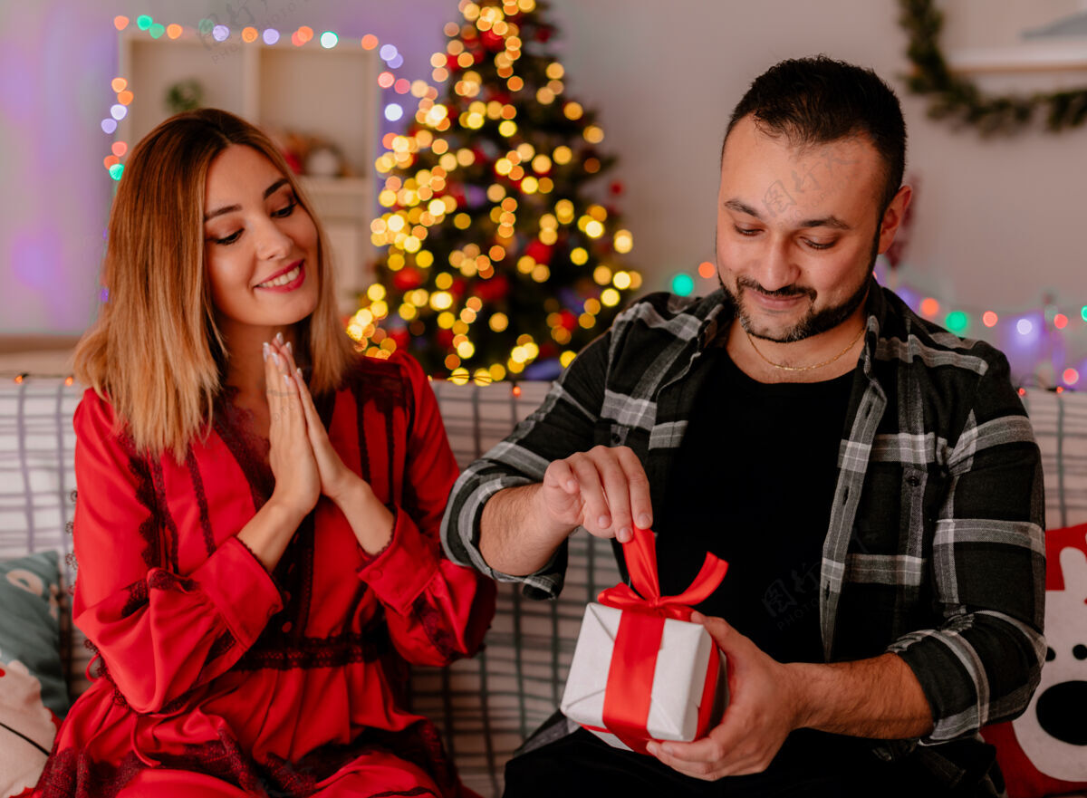 圣诞快乐一对年轻漂亮的情侣坐在沙发上 男人打开礼物 而他的快乐的女友看着他一起庆祝圣诞节在装饰房间圣诞树的背景庆祝圣诞树女朋友