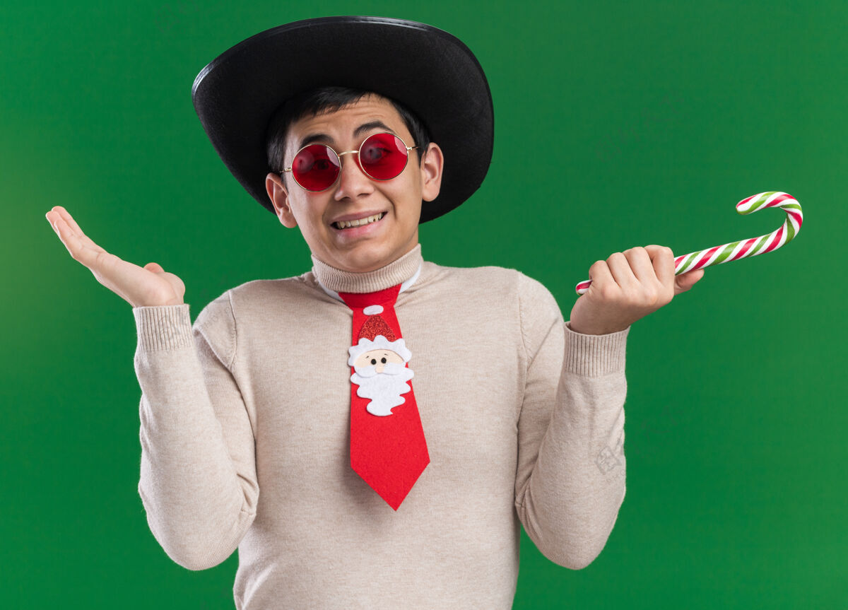领带困惑的年轻人戴着帽子 戴着圣诞领带 戴着眼镜 手里拿着圣诞糖果 双手被隔离在绿色的墙上糖果年轻困惑