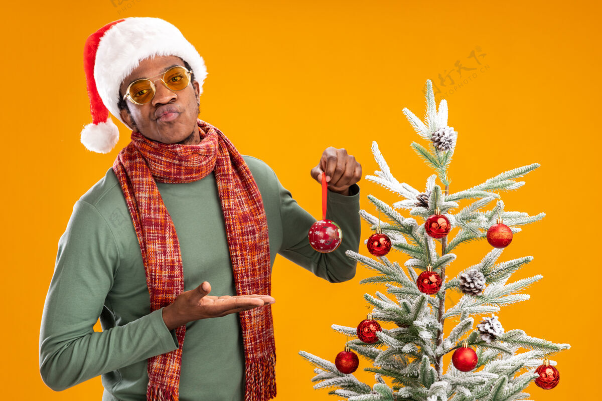 球戴着圣诞帽 围着围巾 脖子上拿着球的非裔美国人站在橙色背景下的圣诞树旁 看起来很不高兴呈现脖子帽子