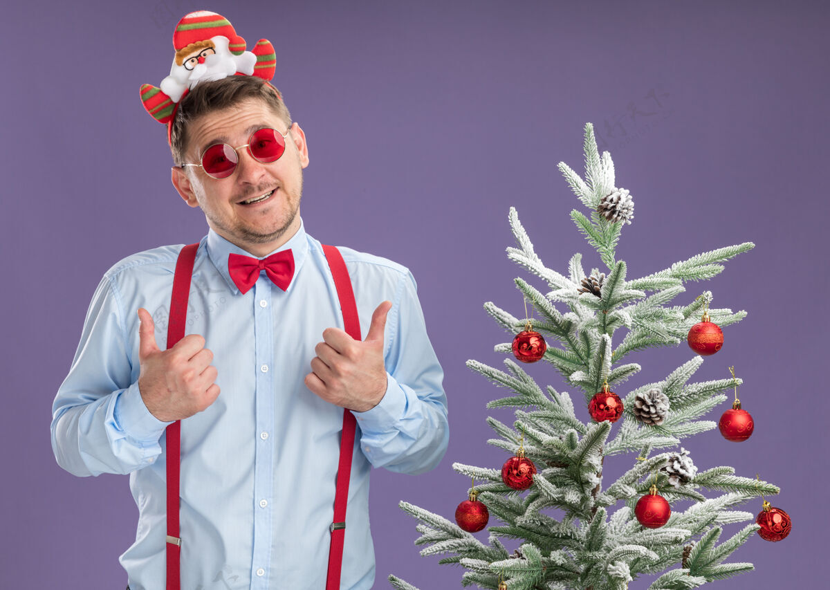 男人快乐的年轻人穿着吊带领结 戴着圣诞老人和红眼镜 站在圣诞树旁微笑着竖起大拇指越过紫色的墙壁吊带站立蝴蝶结