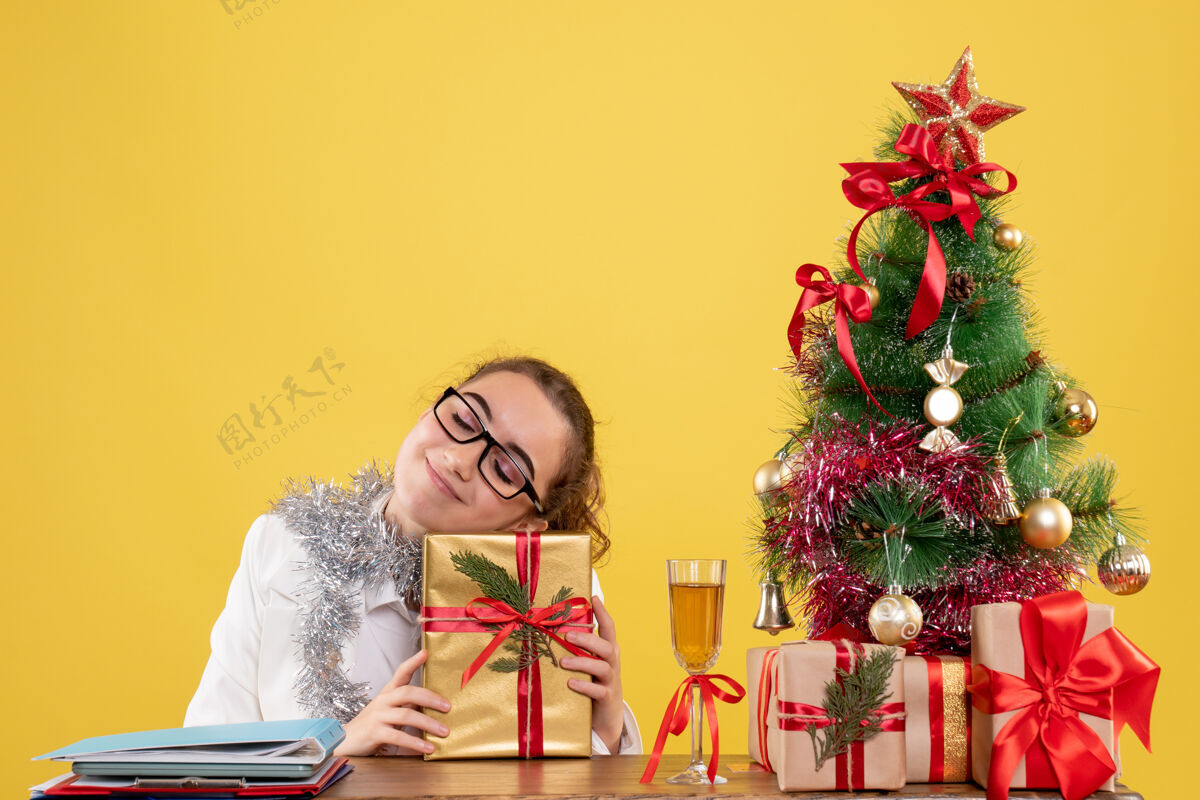 礼物正面图：女医生围坐在圣诞礼物和圣诞树旁 黄色背景上有一张快乐的脸医生丝带高兴