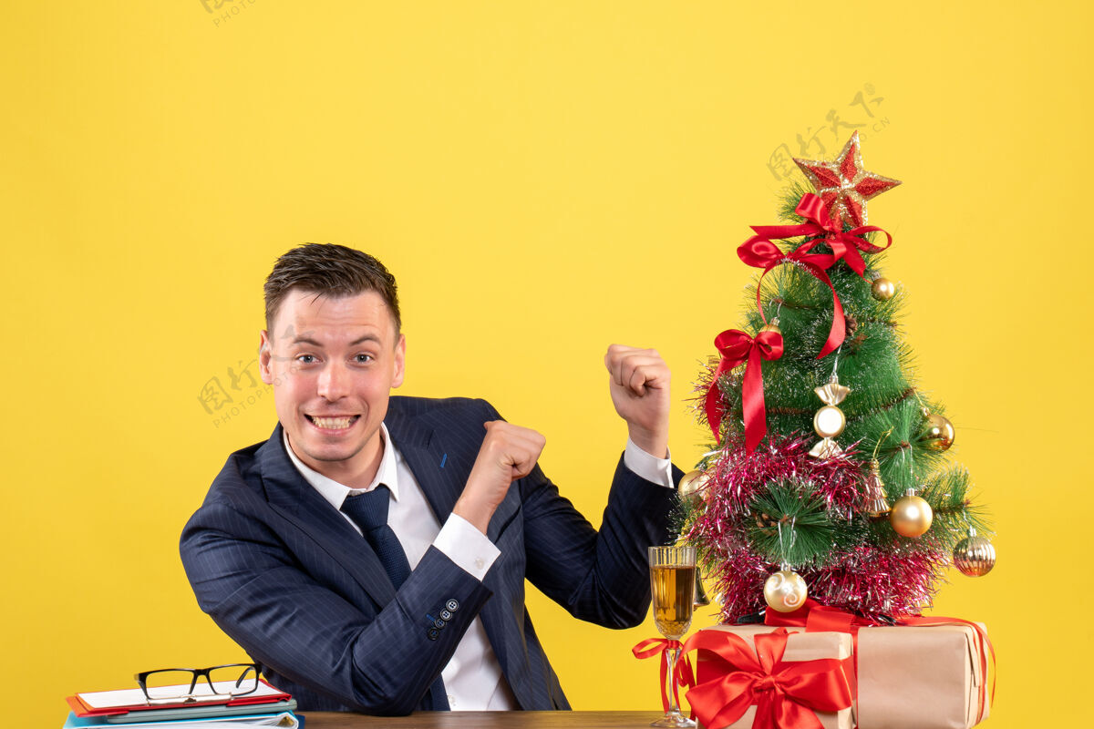办公室正面图是微笑的男人手指着后面坐在圣诞树旁的桌子和黄色的礼物微笑圣诞节手指