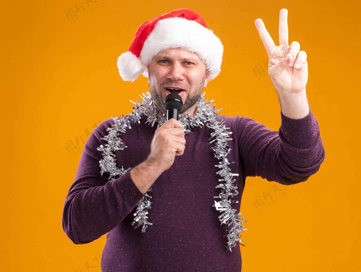 和平自信的中年男子戴着圣诞帽 脖子上戴着金箔花环 对着麦克风说话 在橙色的墙上做着和平的手势圣诞节中年金属片