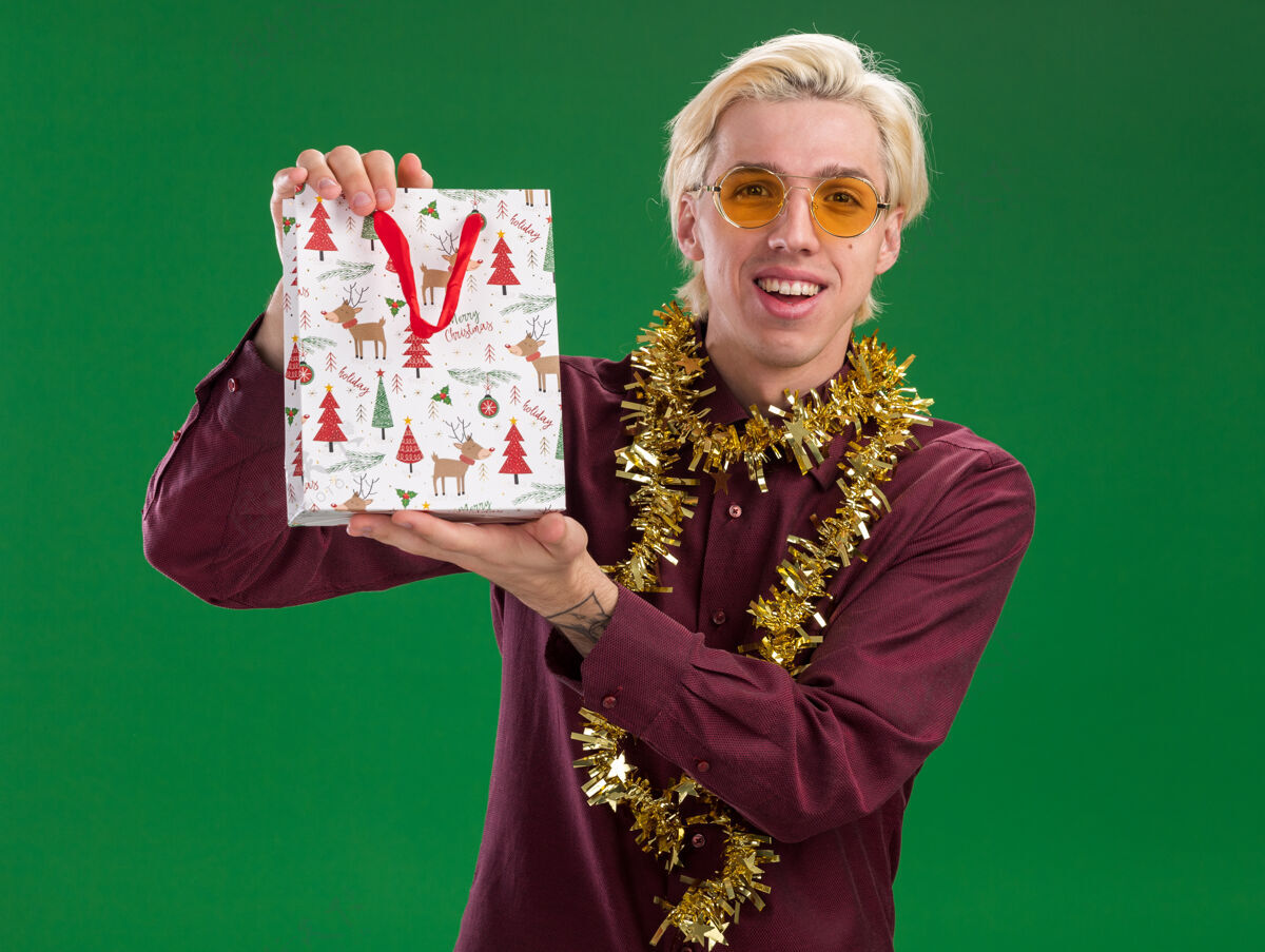 圣诞节快乐的年轻金发男子戴着眼镜 脖子上戴着金箔花环 手里拿着圣诞礼品袋 隔离在绿色的墙上年轻男人穿着