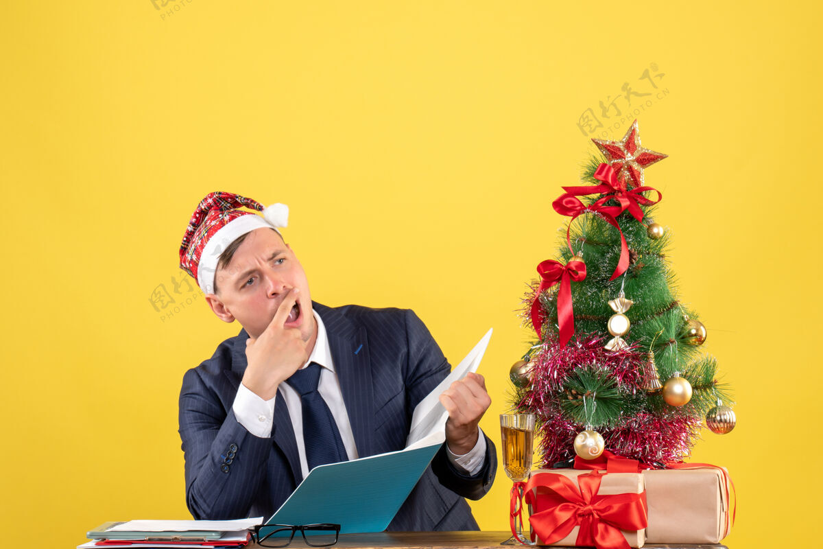 圣诞节困惑的商人坐在圣诞树旁的桌子前 呈现黄色人电脑商人