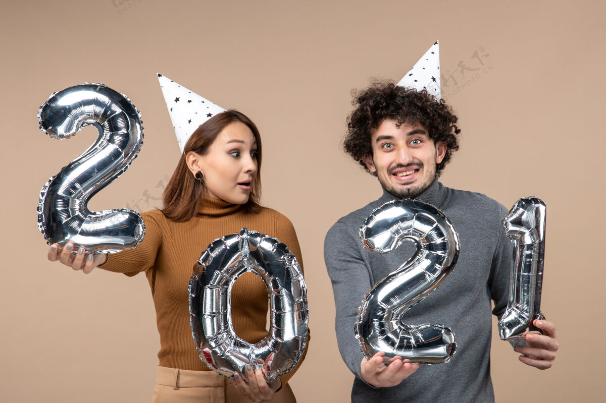 帽子有趣的年轻夫妇戴新年帽的姿势 为相机女孩显示和男子与灰色漂亮模型黄铜