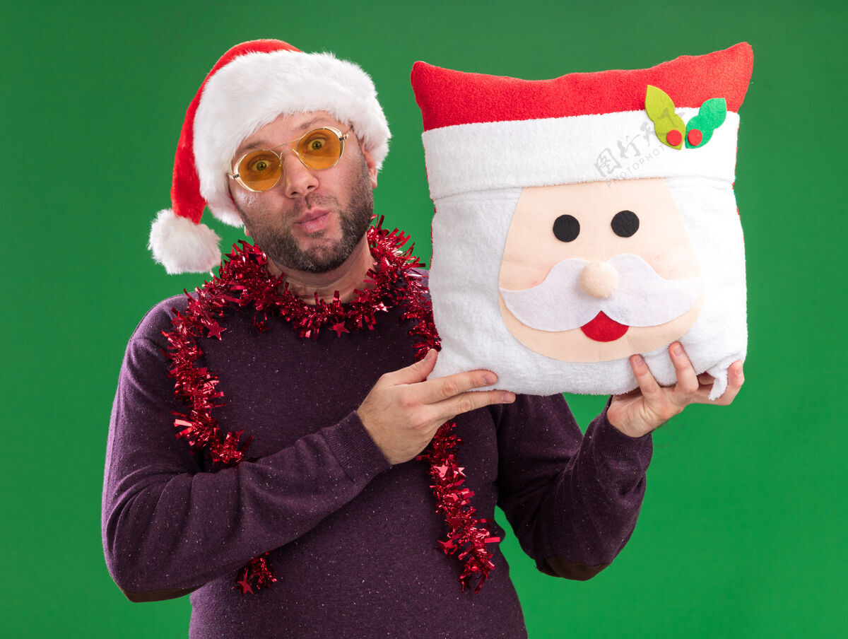 金属丝令人印象深刻的中年男子戴着圣诞帽 脖子上戴着金箔花环 戴着眼镜 抱着圣诞老人的枕头 噘着嘴唇隔离在绿色的墙上花环穿着周围