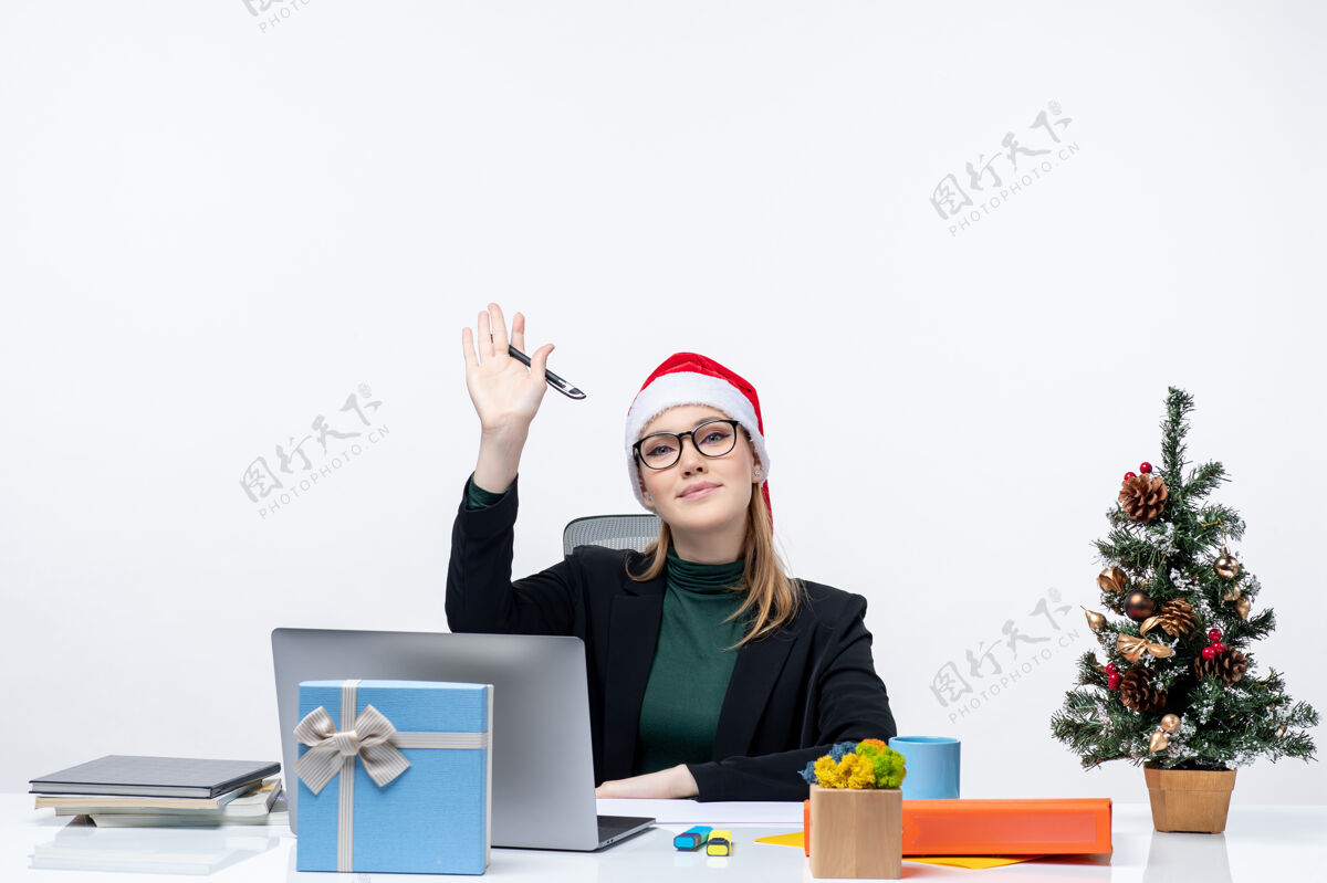 圣诞老人戴着圣诞老人帽的金发女人坐在桌子旁 手里拿着圣诞树和礼物工作制作手势