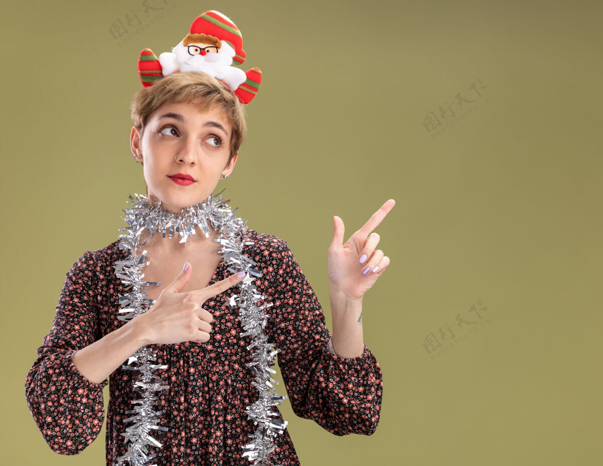 脖子令人印象深刻的年轻漂亮女孩戴着圣诞老人的头带和镀金的花环在脖子上看 并指着橄榄绿墙上的复制空间孤立的一面圣诞老人金属片橄榄