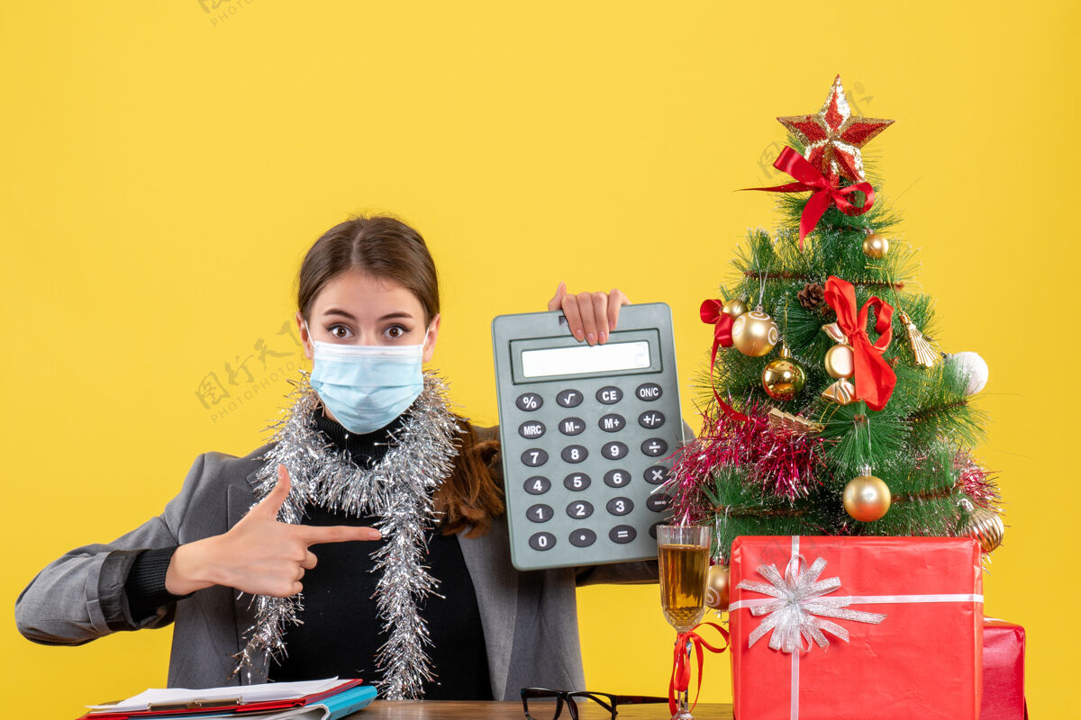 女孩正面图：戴着医用面具的女孩坐在桌子旁 手里拿着计算器圣诞树和鸡尾酒好奇成人鸡尾酒