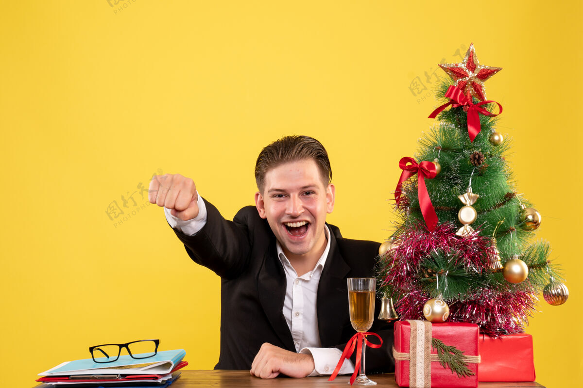 年正面图年轻的男工人坐在圣诞礼物和圣诞树喜悦情感颜色男