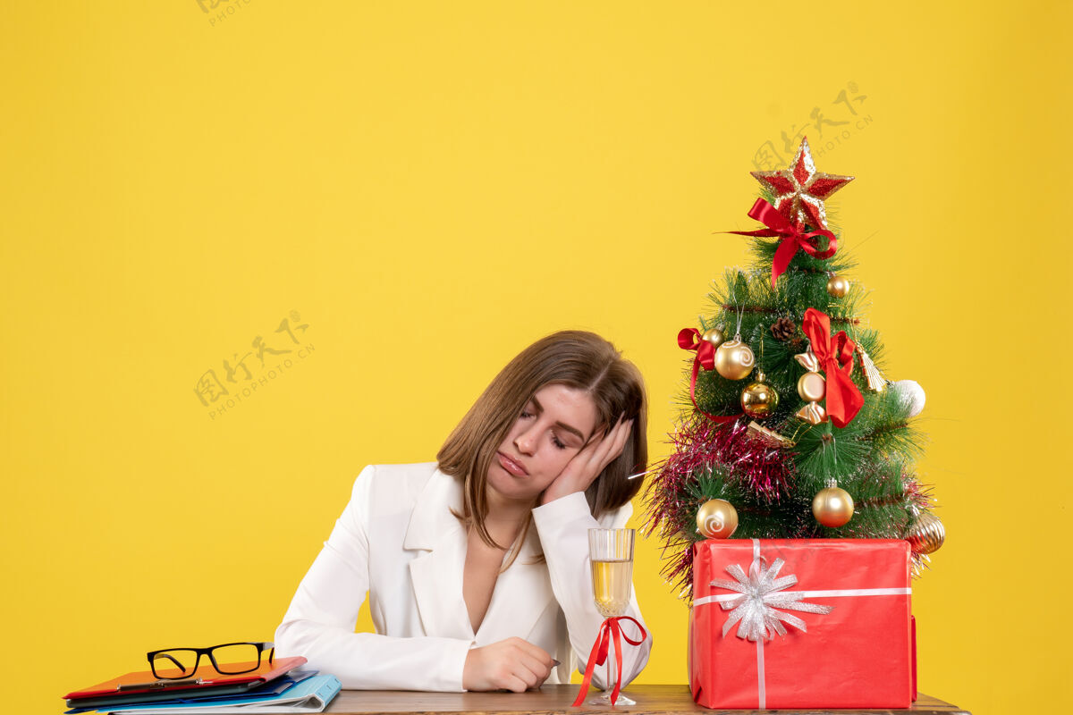 人正面图：女医生坐在黄色背景下疲惫的桌子前 带着圣诞树和礼品盒情绪肖像圣诞节