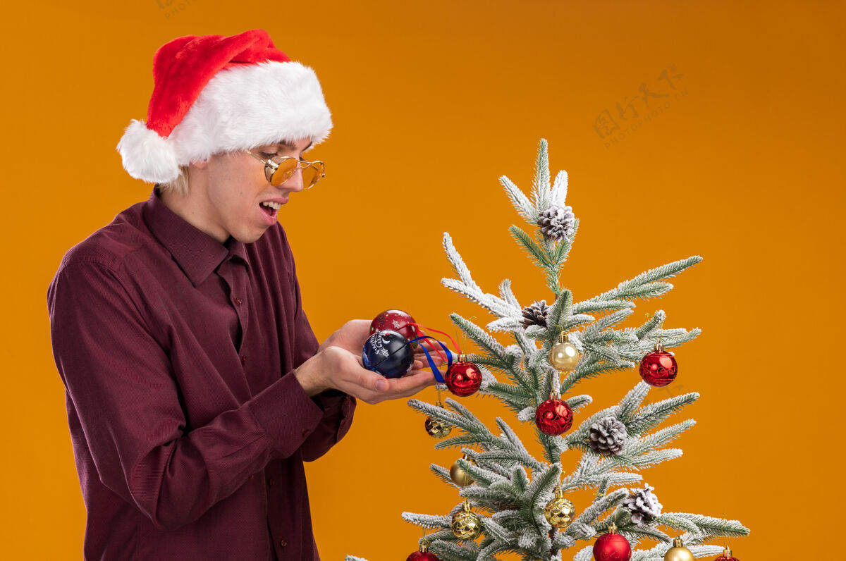 圣诞树令人印象深刻的年轻金发男子戴着圣诞帽和眼镜站在侧面图装饰圣诞树附近的橙色背景圣诞老人金发看