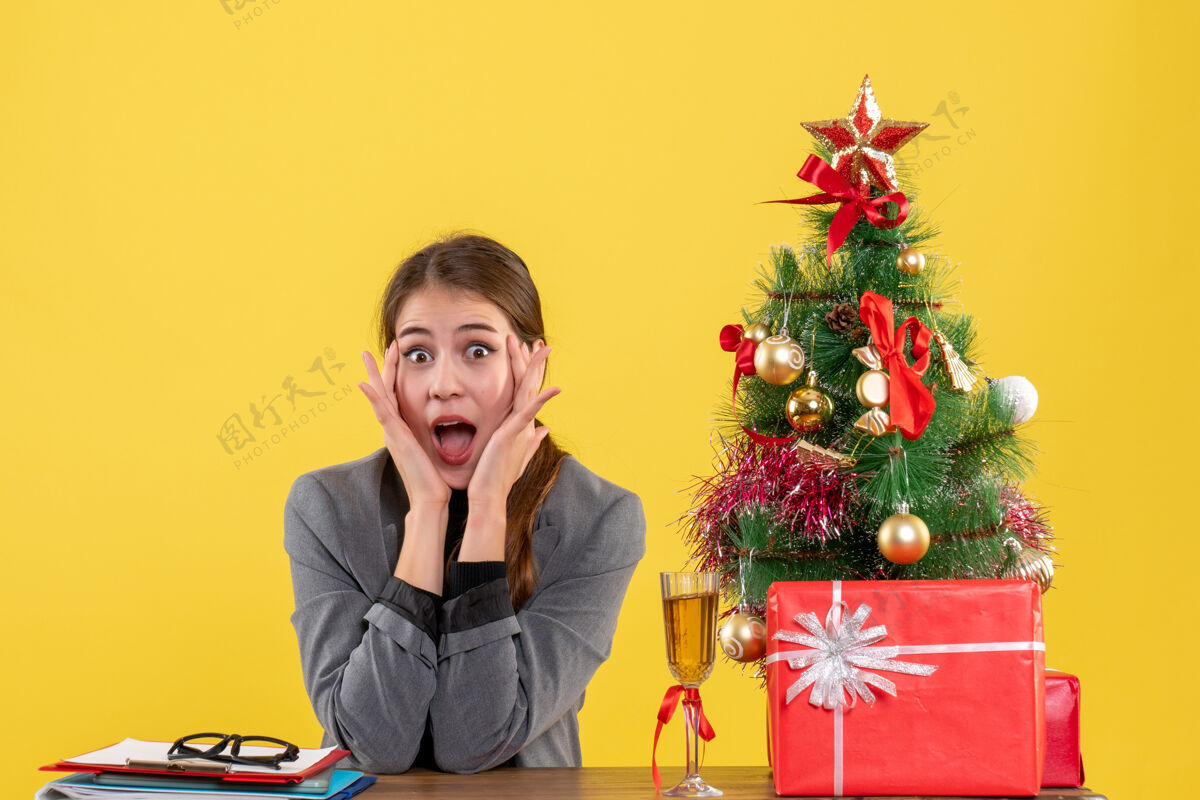 坐着前视图惊讶的女孩坐在桌子旁 把手放在脸颊上 靠近圣诞树和礼物鸡尾酒树女孩手