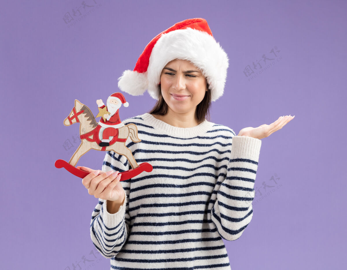 快乐无知的年轻白人女孩拿着圣诞老人的帽子 看着摆在摇马上的圣诞老人圣诞老人女孩帽子