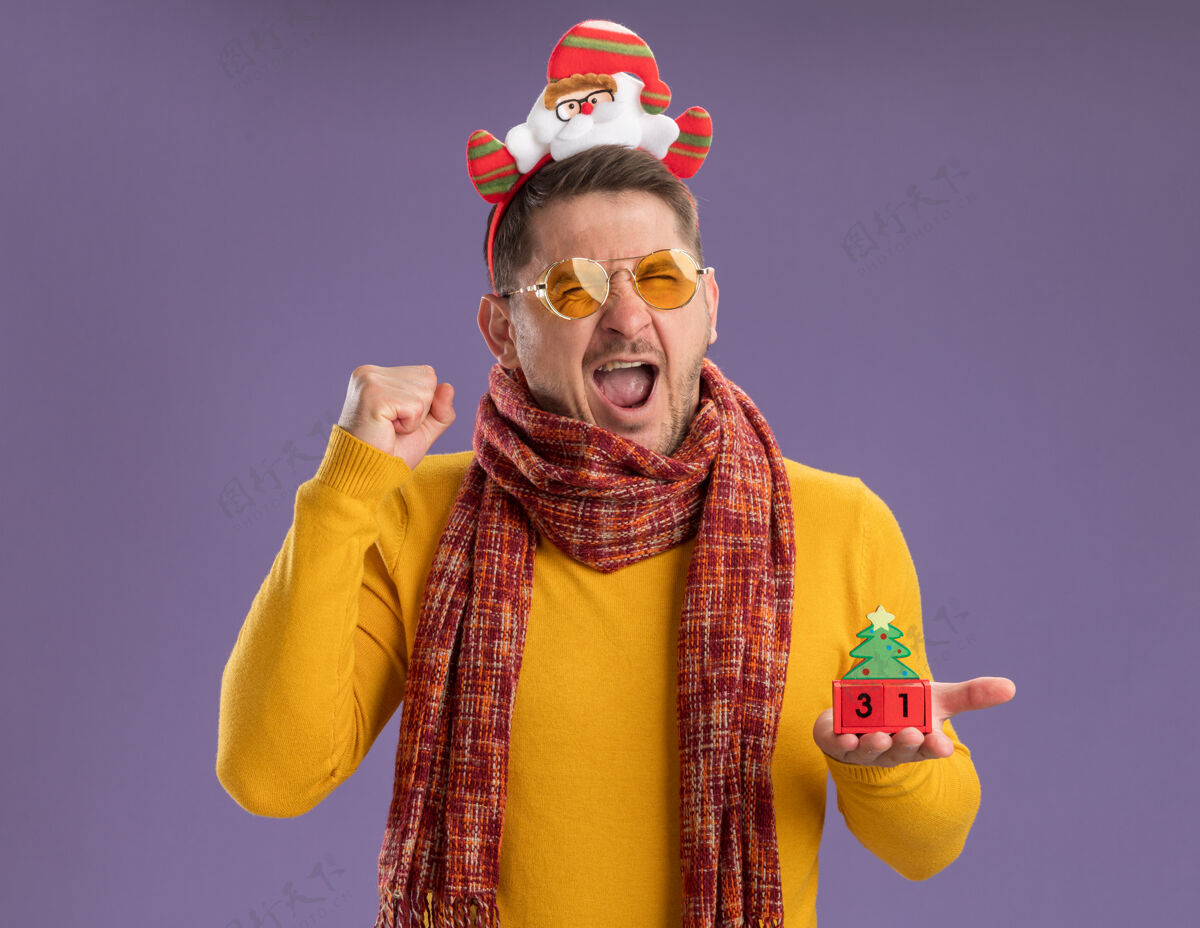 边缘穿着黄色高领毛衣 戴着暖和的围巾和眼镜的年轻人戴着滑稽的帽沿 头上戴着圣诞老人 展示着新年约会的玩具立方体 握紧拳头疯狂快乐地站在紫色的墙上显示日期拳头