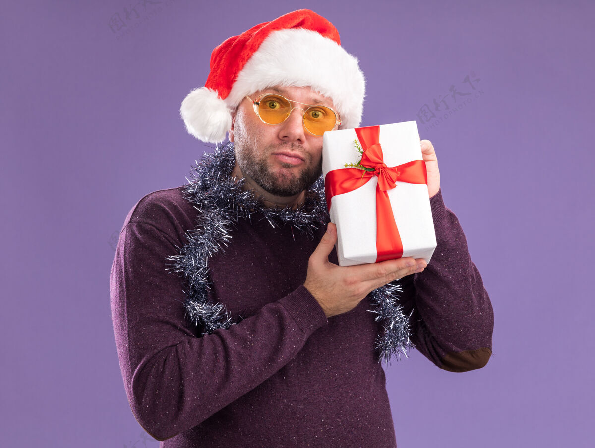 圣诞节困惑的中年男子戴着圣诞帽 脖子上戴着金箔花环 戴着眼镜 手里拿着礼包 隔离在紫色的墙上周围包装中年