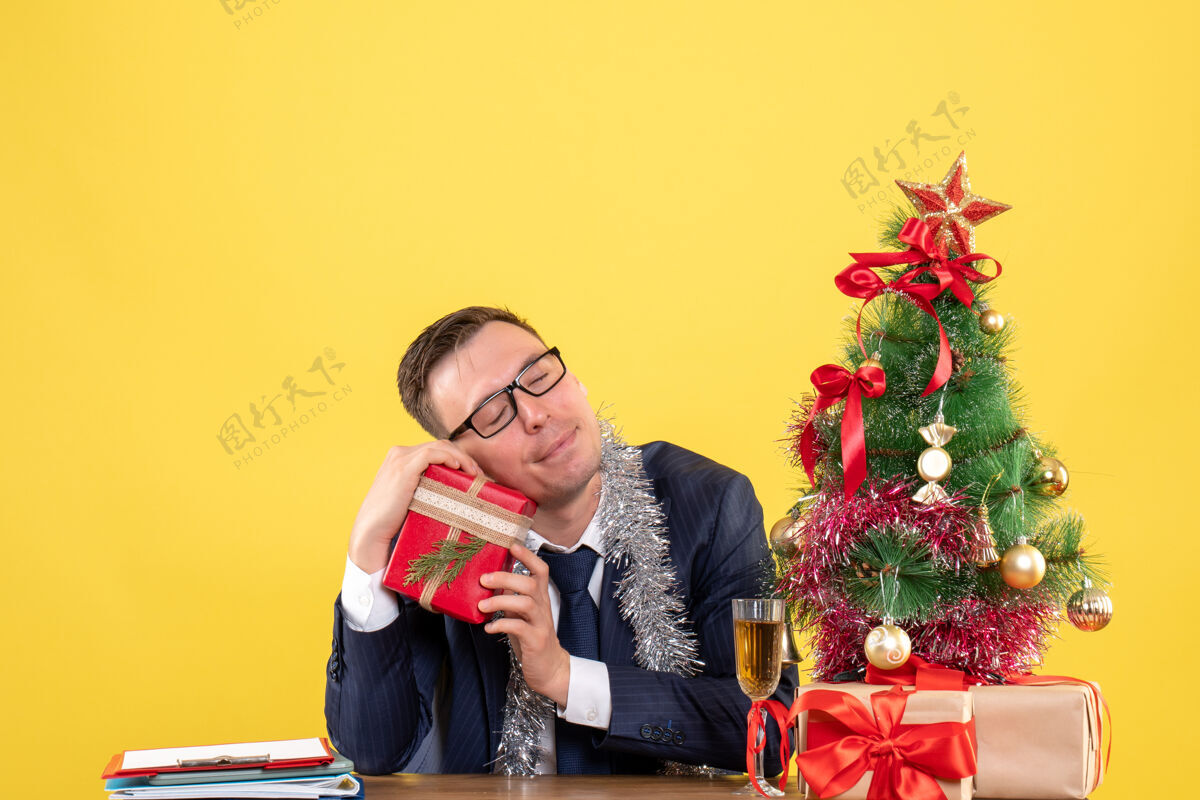 礼物前视图满意的人闭着眼睛拿着他的礼物坐在圣诞树旁边的桌子上 黄色的礼物黄色坐着肖像