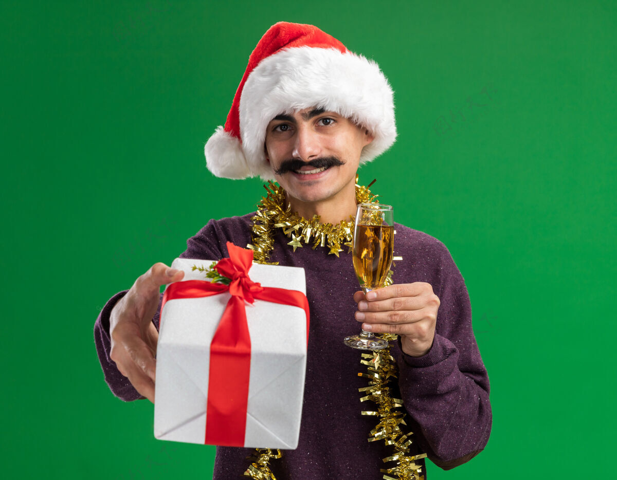 圣诞快乐快乐的小胡子男人戴着圣诞老人的帽子 脖子上戴着金属丝 手里拿着一杯香槟和圣诞礼物 站在绿色的背景下 面带微笑地看着相机香槟快乐金属片