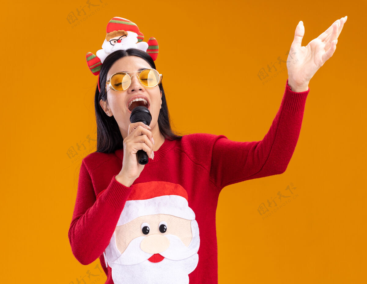 毛衣年轻的白人女孩戴着圣诞老人的头带和毛衣戴着眼镜拿着麦克风近嘴唱歌闭着眼睛保持手在空气中隔离橙色背景手圣诞老人嘴