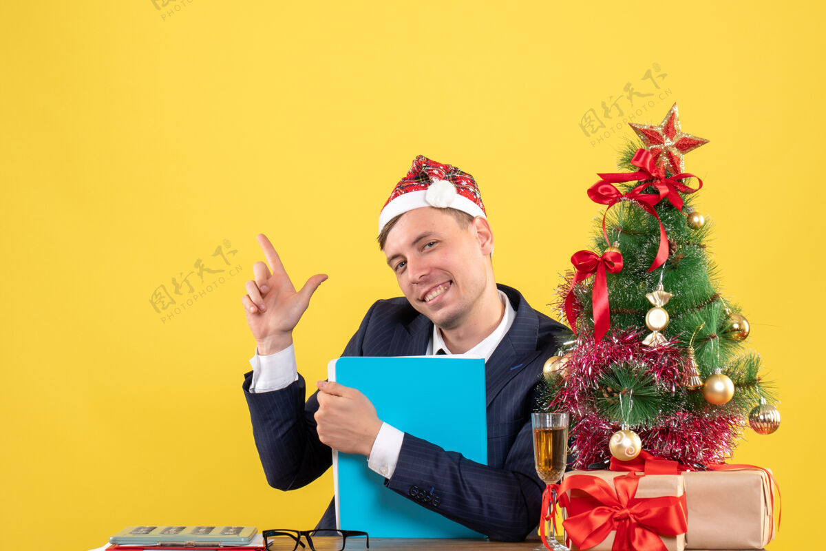 桌子前视图幸福的商人拿着文件文件坐在圣诞树附近的桌子上 黄色的礼物抱着成人文件