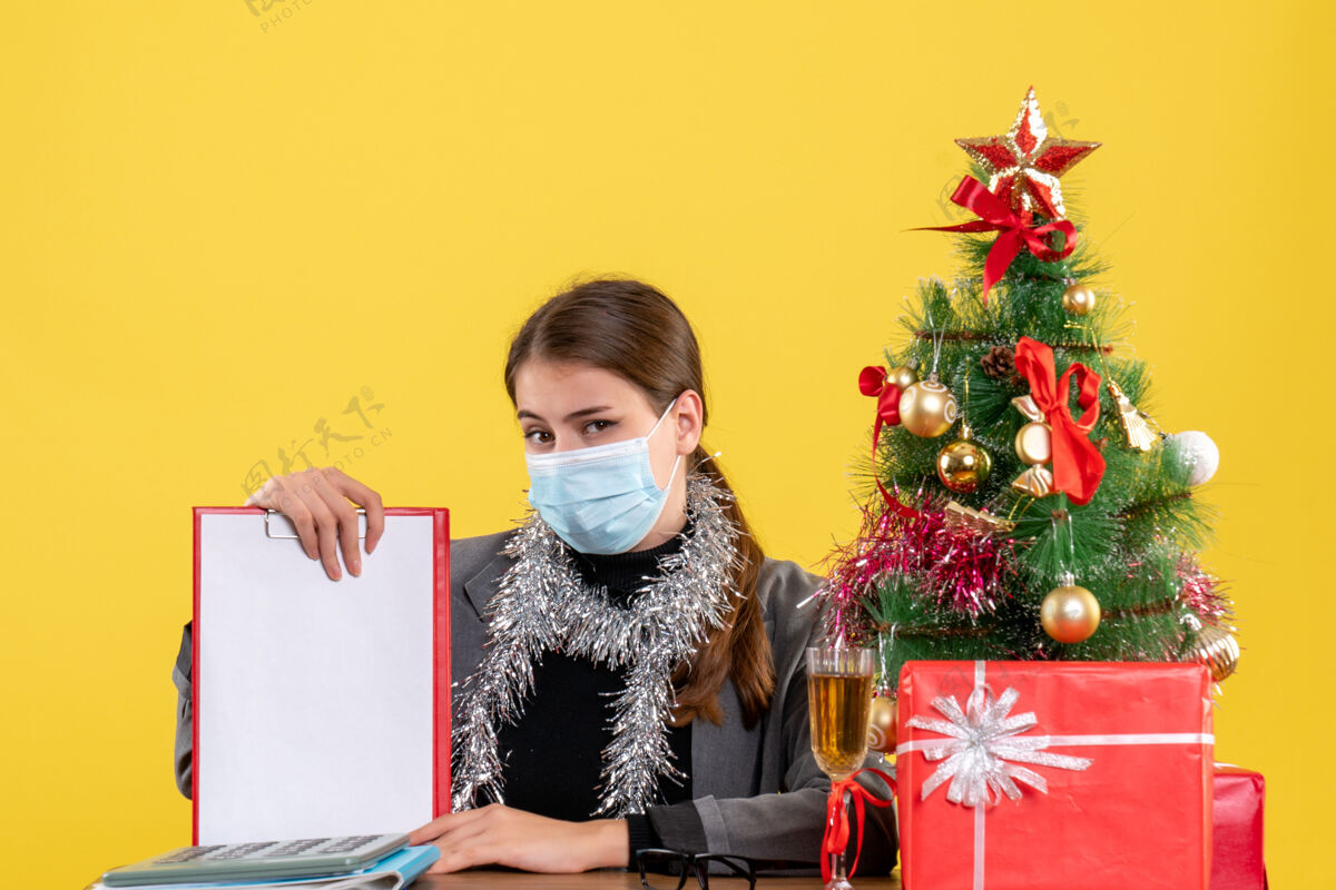 女孩正面图：戴着医用面具的坚强女孩坐在圣诞树和鸡尾酒礼品的桌边圣诞节鸡尾酒面具