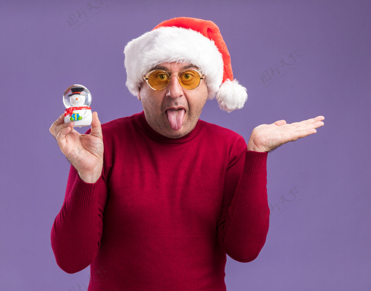 眼镜滑稽的中年男子戴着圣诞帽戴着黄色眼镜拿着圣诞雪球伸出舌头举起胳膊站在紫色背景上圣诞节中间舌头