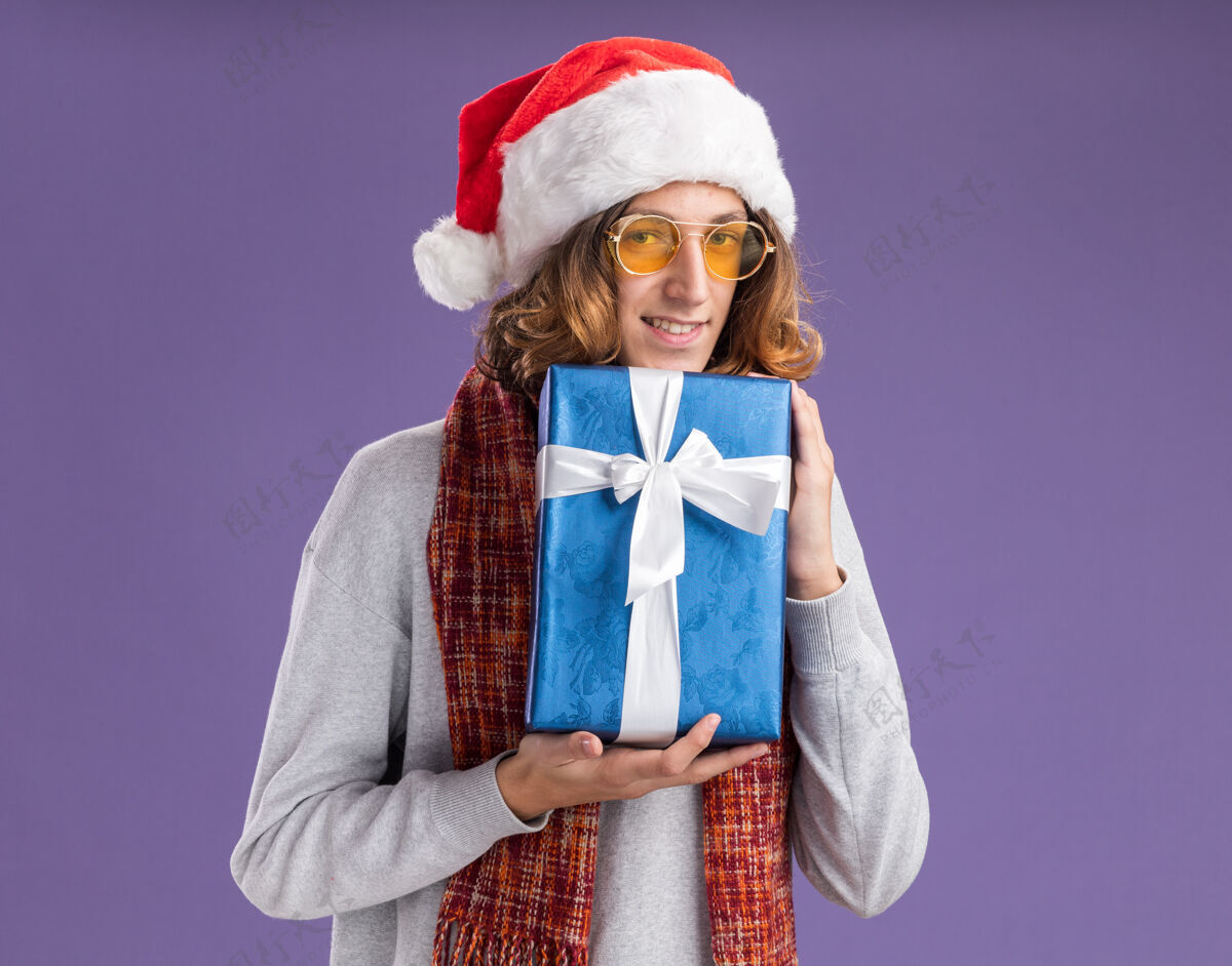 圣诞节快乐的年轻人戴着圣诞老人帽 戴着黄色眼镜 脖子上围着暖和的围巾 手里拿着圣诞礼物 站在紫色的背景下 看着相机 兴高采烈地笑着礼物圣诞老人围巾