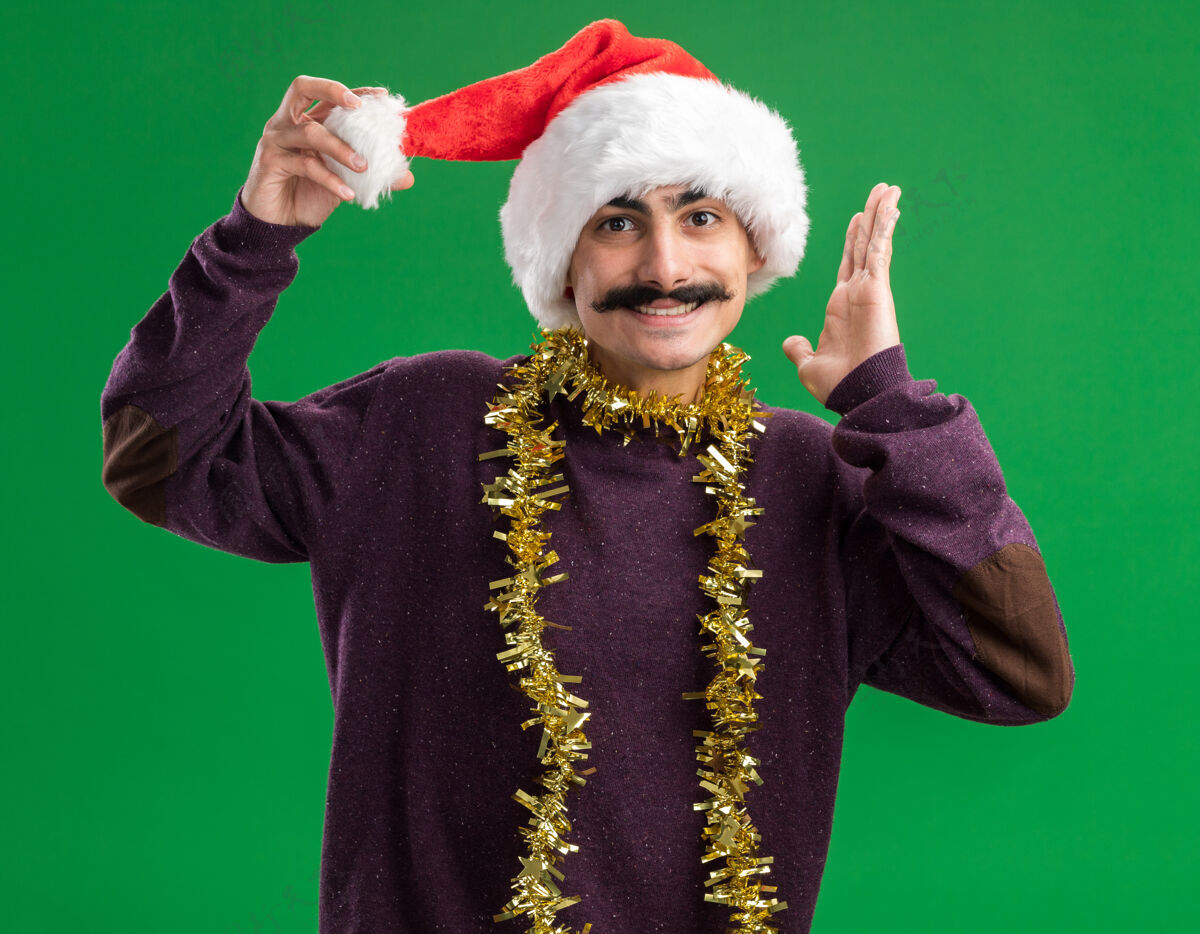 金属片年轻的胡子男人戴着圣诞老人帽子 脖子上戴着金属丝 站在绿色的背景下 看着相机 满脸笑容年轻脸脖子